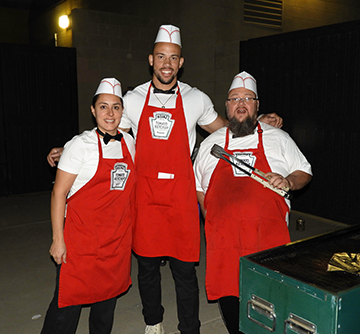 Three staff members wearing Heinz Tomato Ketchup aprons and food preparation hats pose together beside a grill