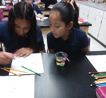 A female student looking over another female student's shoulder as they work on a classroom project with colored pencils together