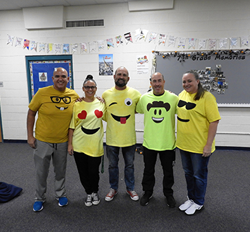 Five staff members wearing emoji t-shirts pose together in a classroom
